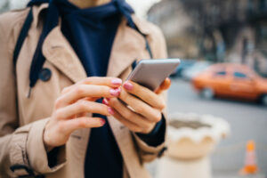 Young woman wearing beige coat using smart phone standing near road with cars in city at spring day. 